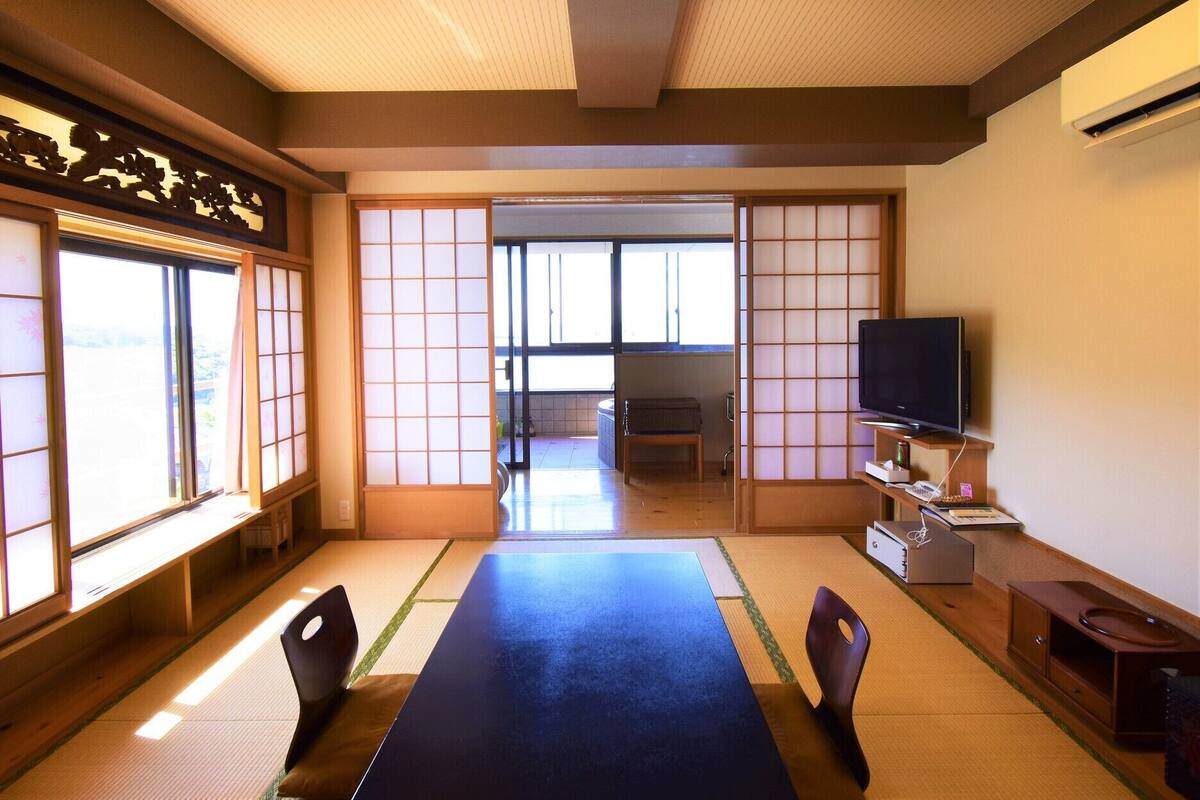 Ein traditioneller japanischer Raum mit Tatami-Böden, einem niedrigen Tisch, Stühlen, einem Fernseher und Schiebetüren.