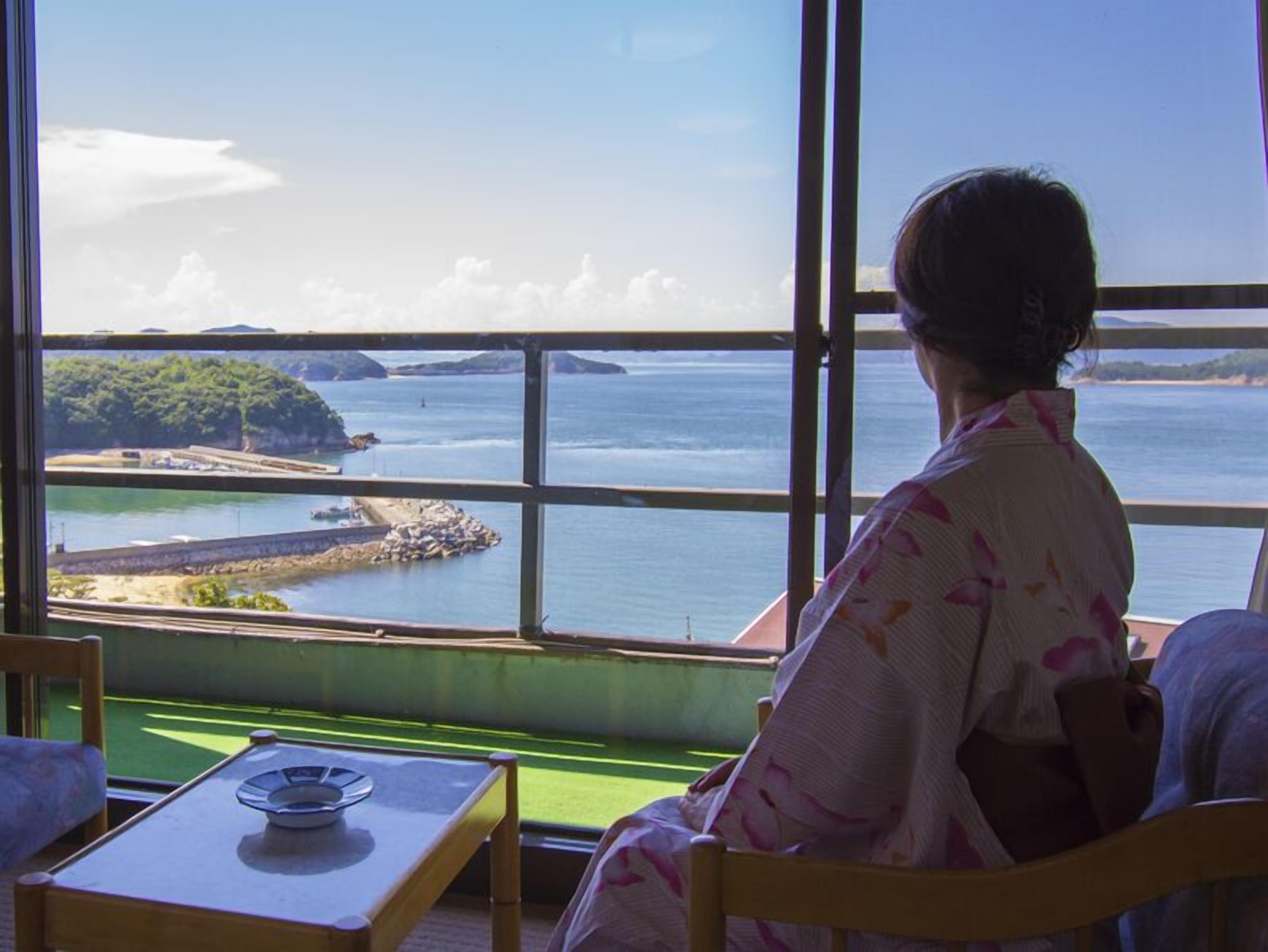 A room with a large window overlooking a coastal area, a table with a plate, and a chair.