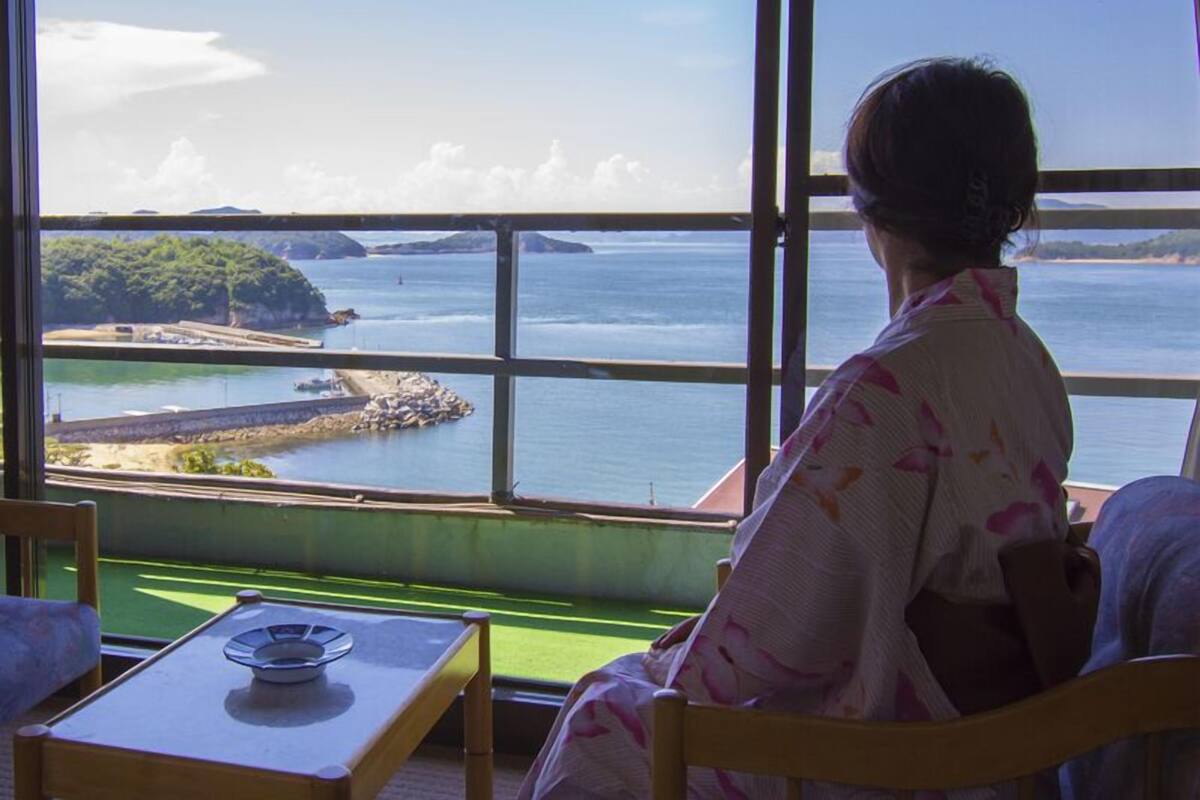 Ein Zimmer mit einem großen Fenster mit Blick auf eine Küstenlandschaft, einem Tisch mit Teller und einem Stuhl.