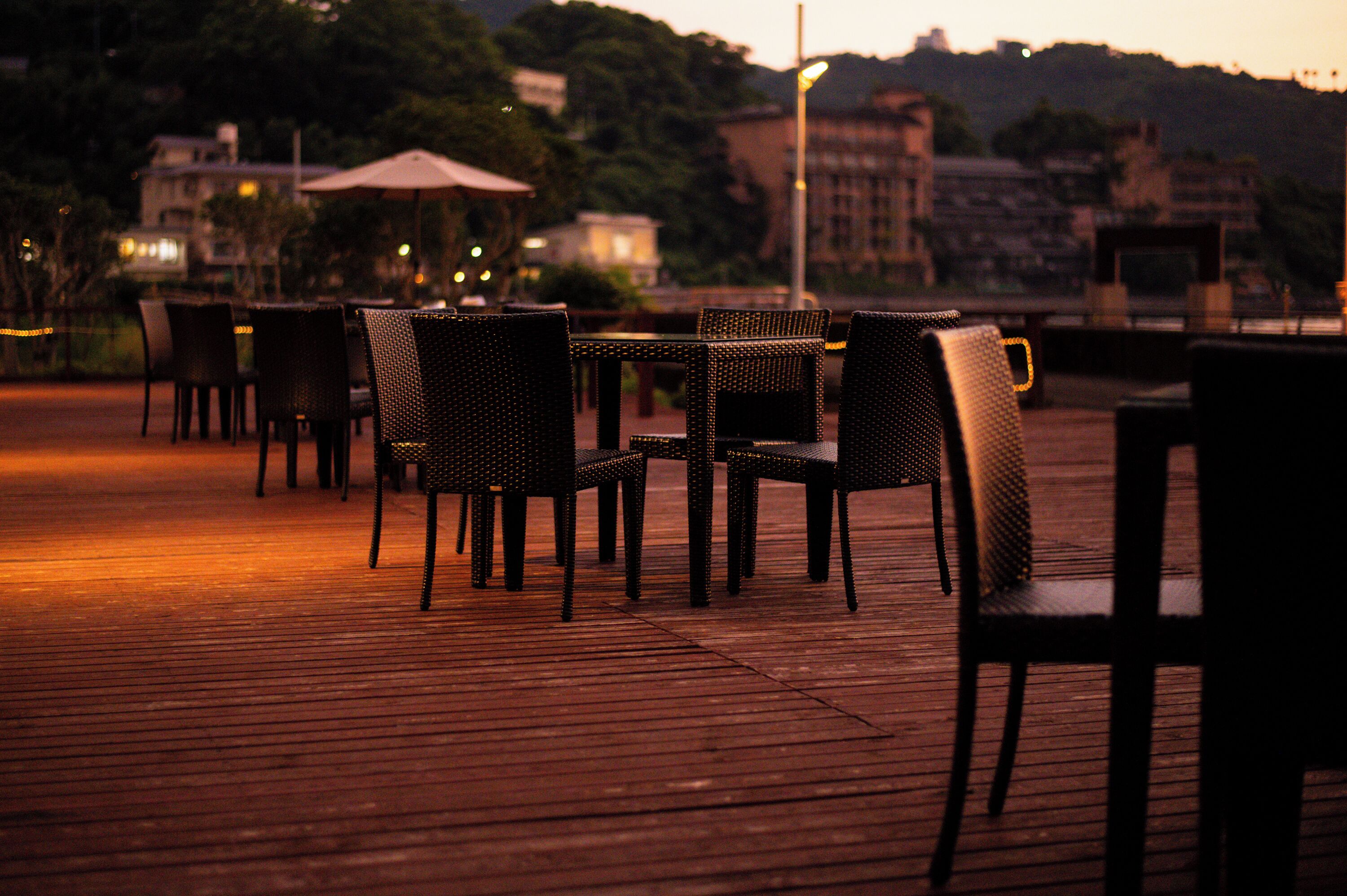 An outdoor dining area with wicker chairs and tables, surrounded by a wooden deck and overlooking a cityscape.
