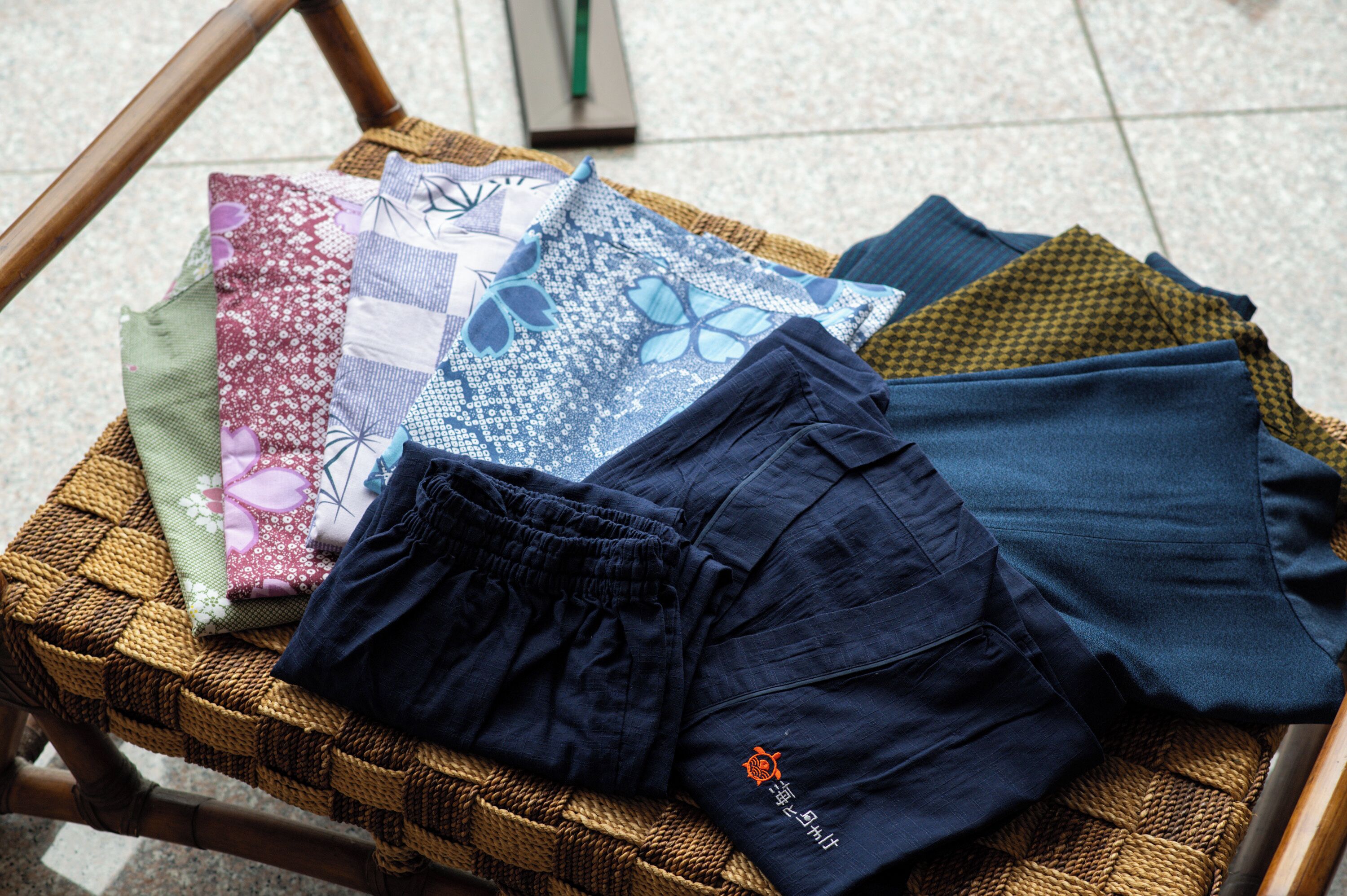 Een gevlochten mand met verschillende stoffen en een broek van donkerblauw op een betegelde vloer.