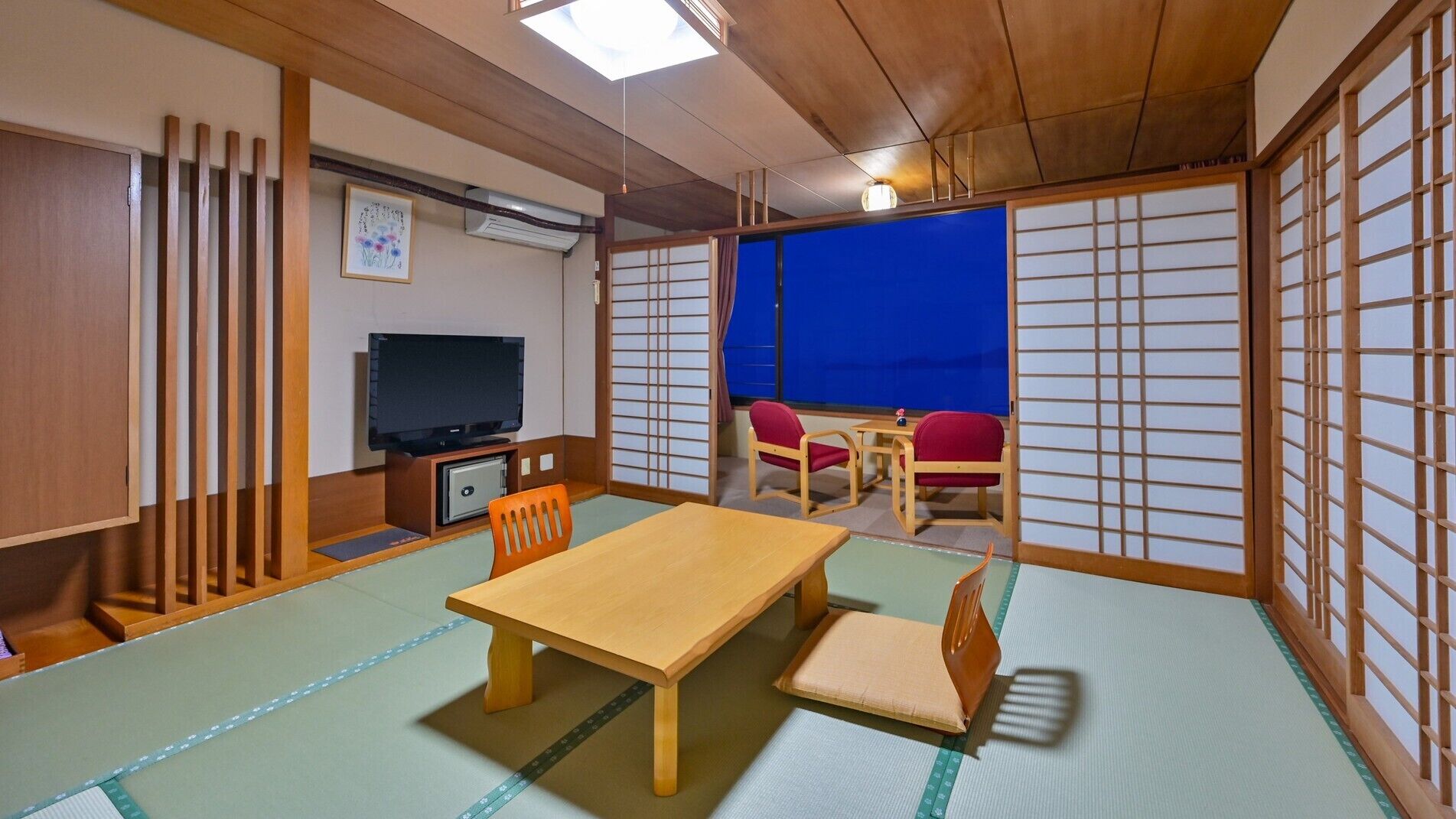 A tatami mat room with a wooden table, chairs, a TV, and a view through shoji screens.
