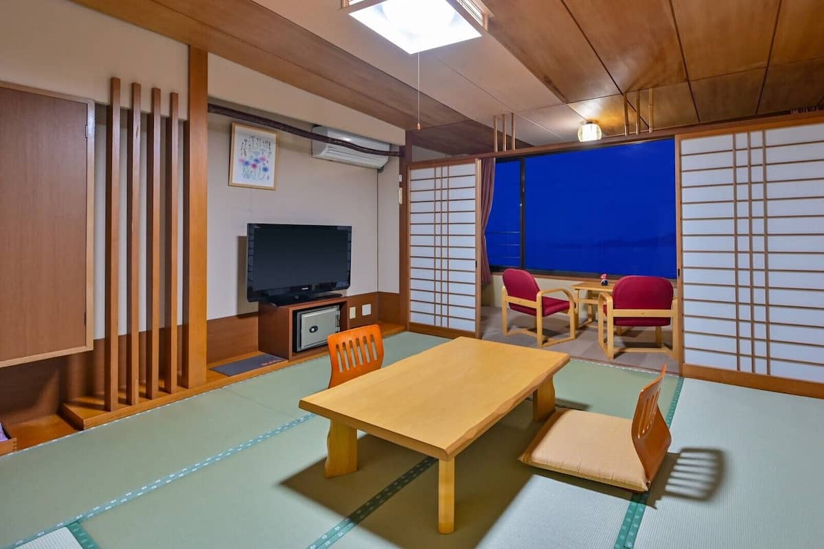 A tatami mat room with a wooden table, chairs, a TV, and a view through shoji screens.