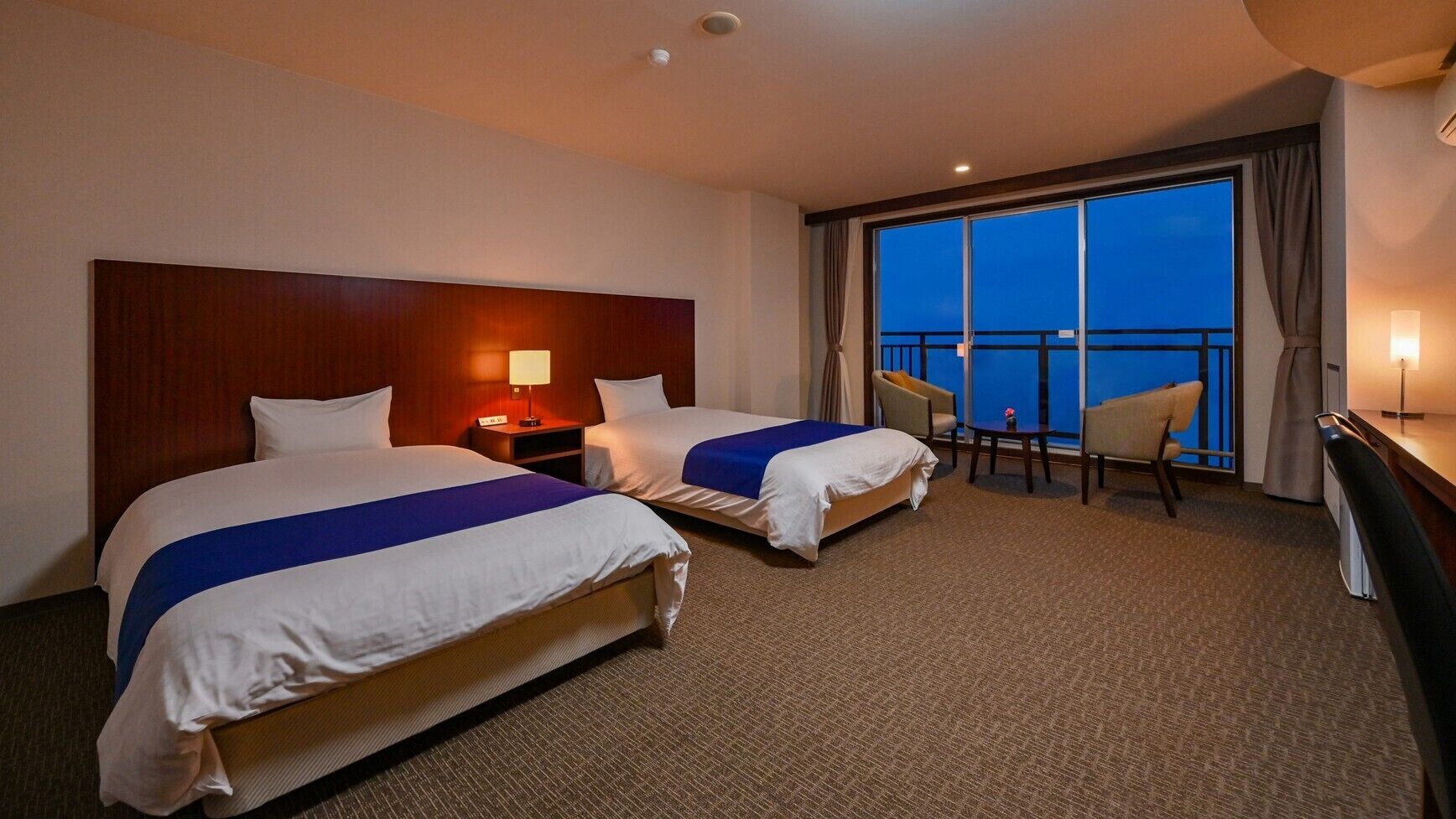 A hotel room with two beds, a desk, a chair, a nightstand, and a large window with a view.