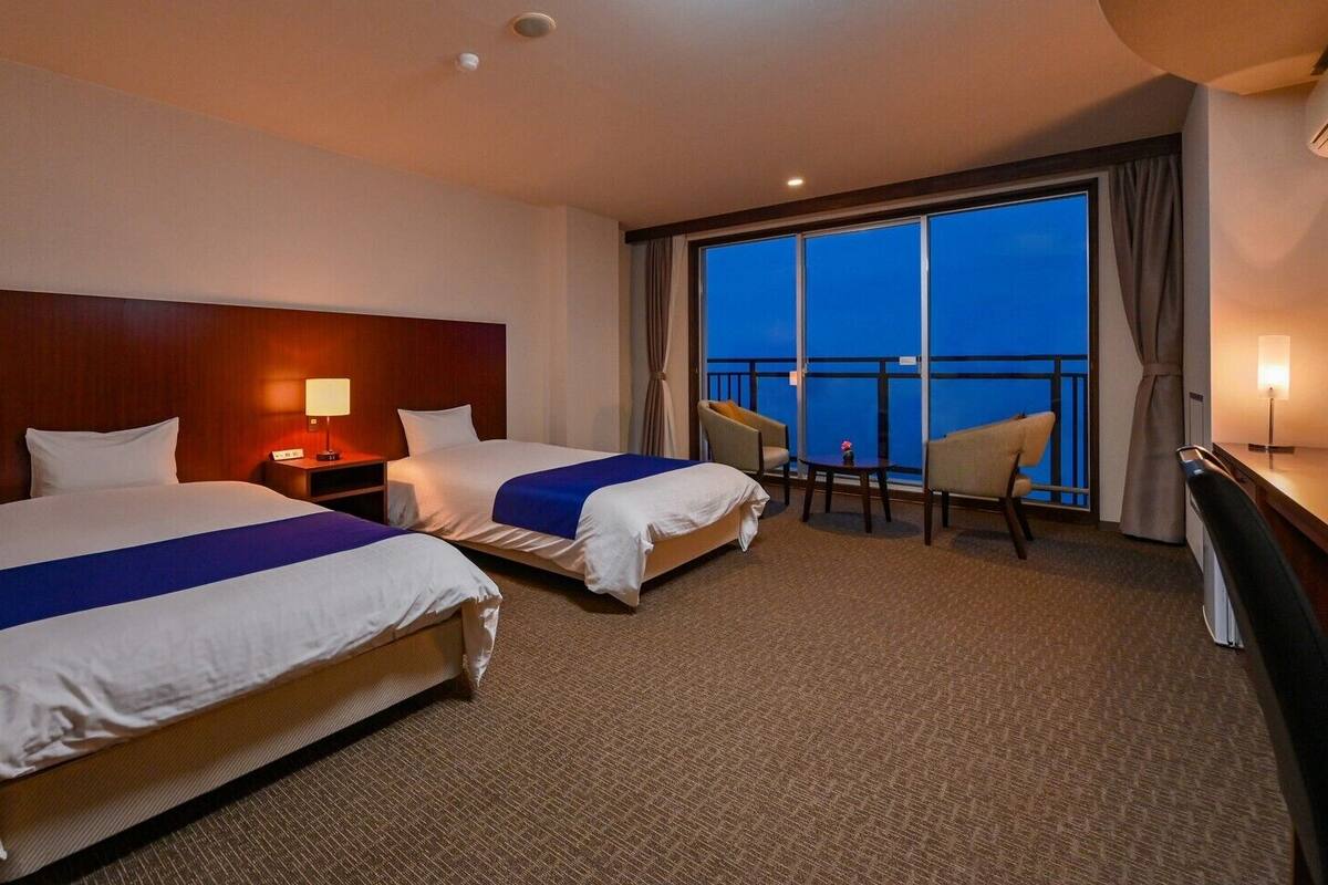 A hotel room with two beds, a desk, a chair, a nightstand, and a large window with a view.