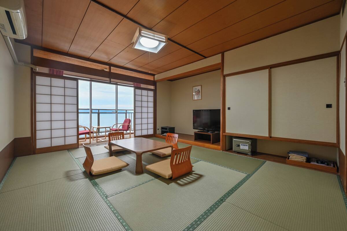 A traditional Japanese room with tatami flooring, a low table, chairs, a television, and sliding doors leading to a balcony with a view of the sea.