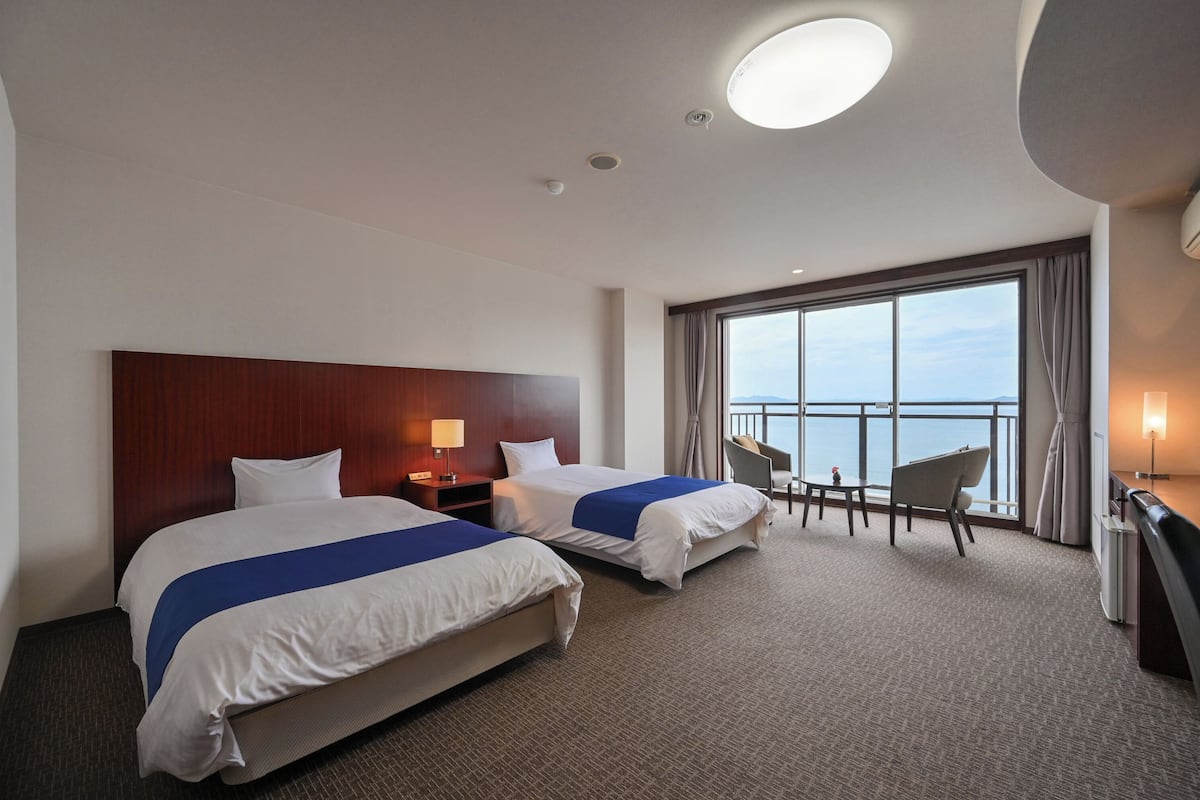 A hotel room with two beds, a desk, a chair, and a large window with a view of the sea.
