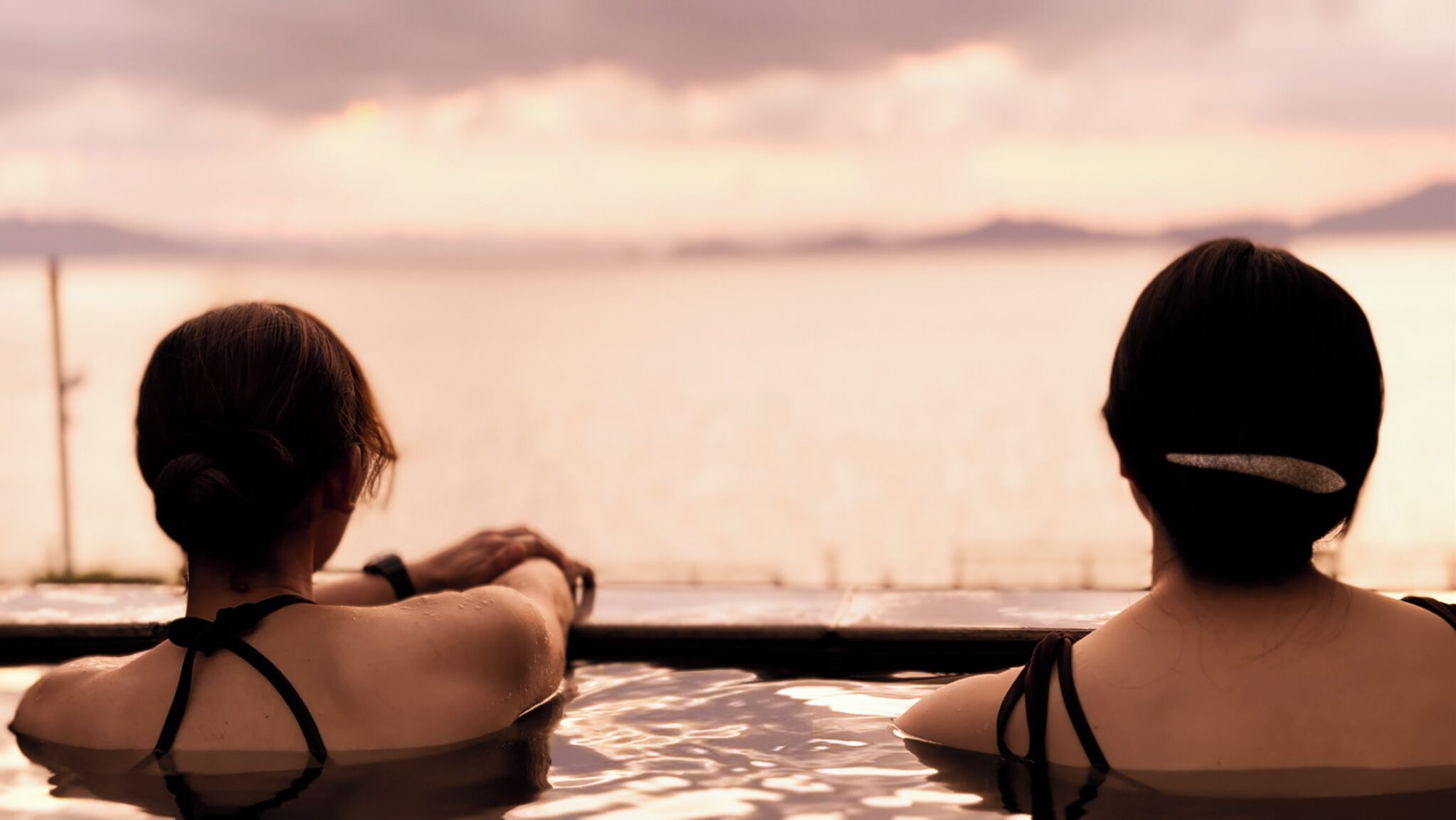 Two individuals seated by a poolside, overlooking a body of water with a distant shoreline.