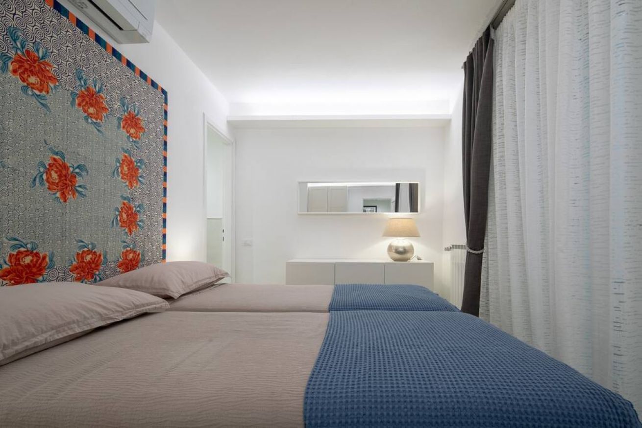 A hotel room with two beds, a bedside table with a lamp, a mirror, and a window with curtains.