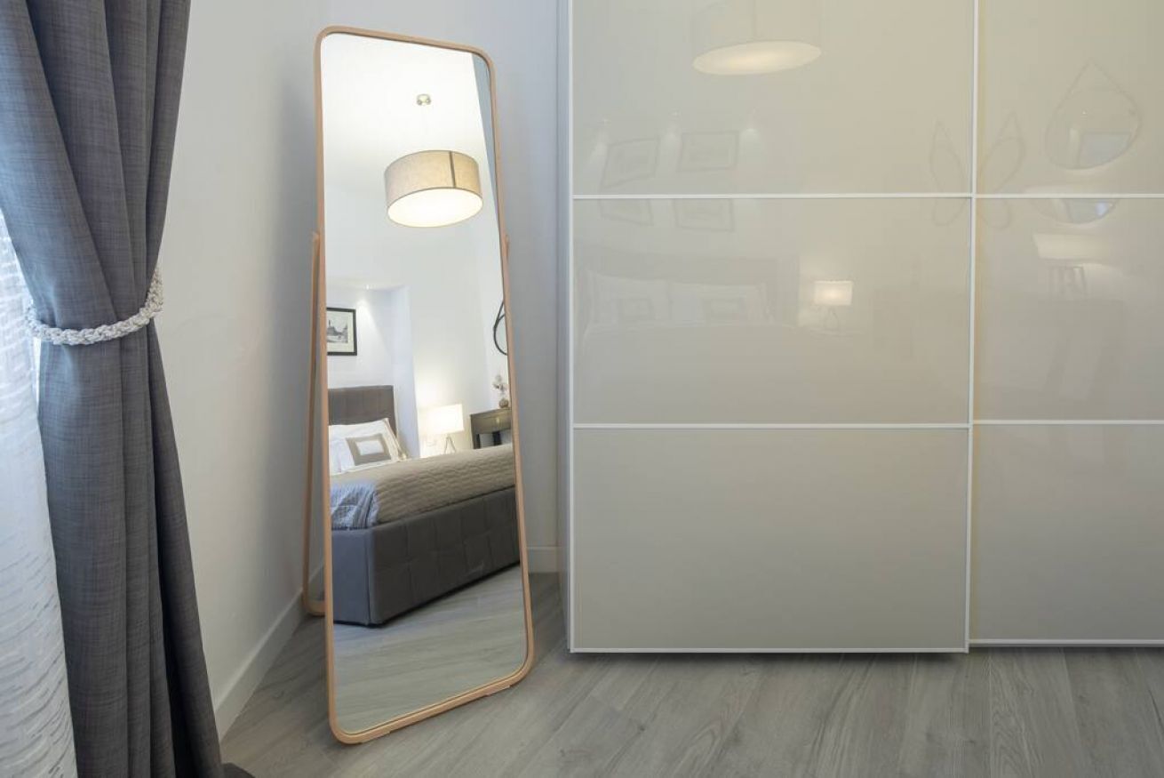 A room with a large mirror, a bed, a bedside table, and a wall with a light fixture.