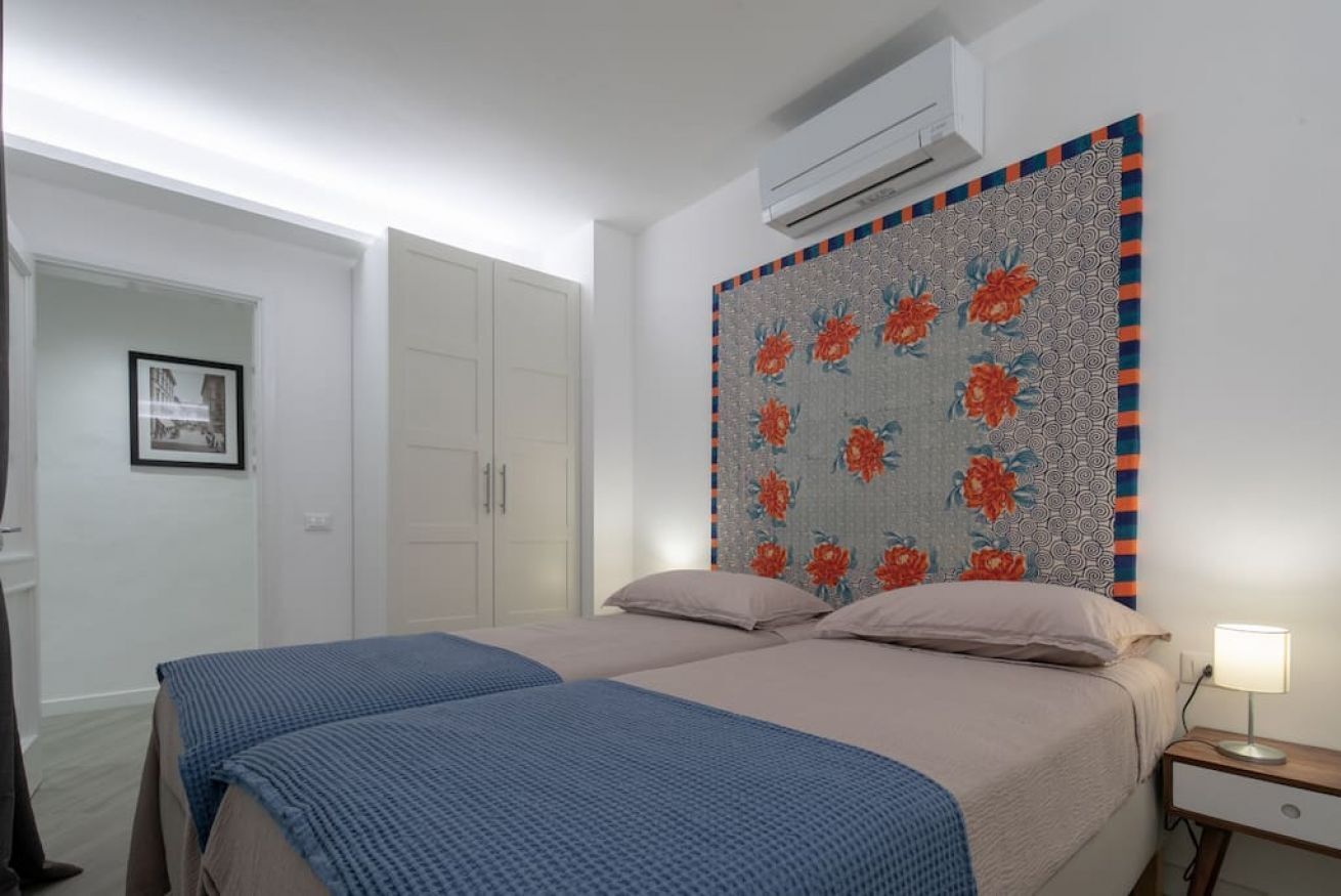Et soveværelse med to senge, en stor sengegavl med blomstermønster, et hvidt skab og en aircondition-enhed.
