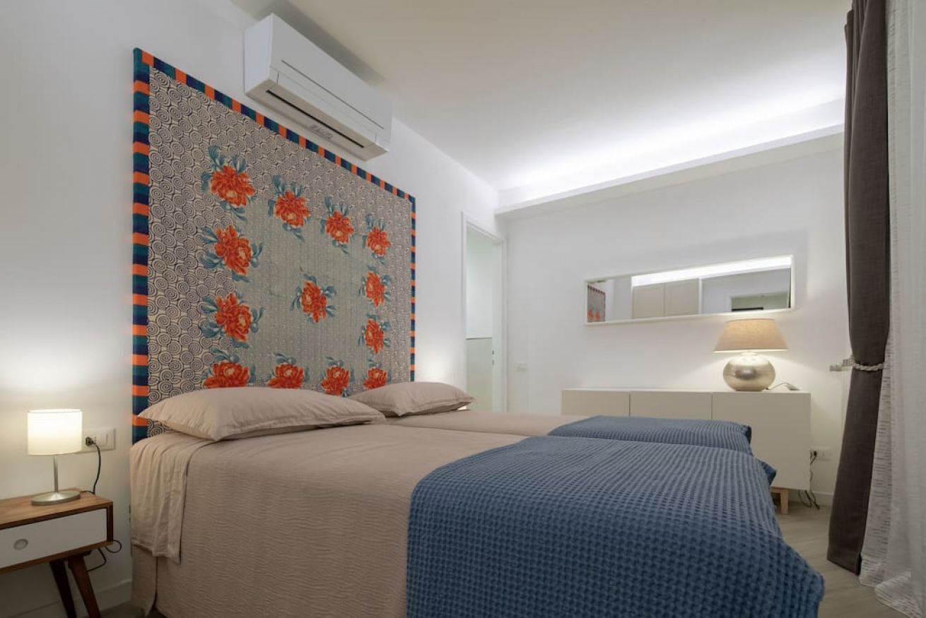 A bedroom with two beds, a bedside table with a lamp, a large floral-patterned headboard, a mirror above the bed, and a window with curtains.