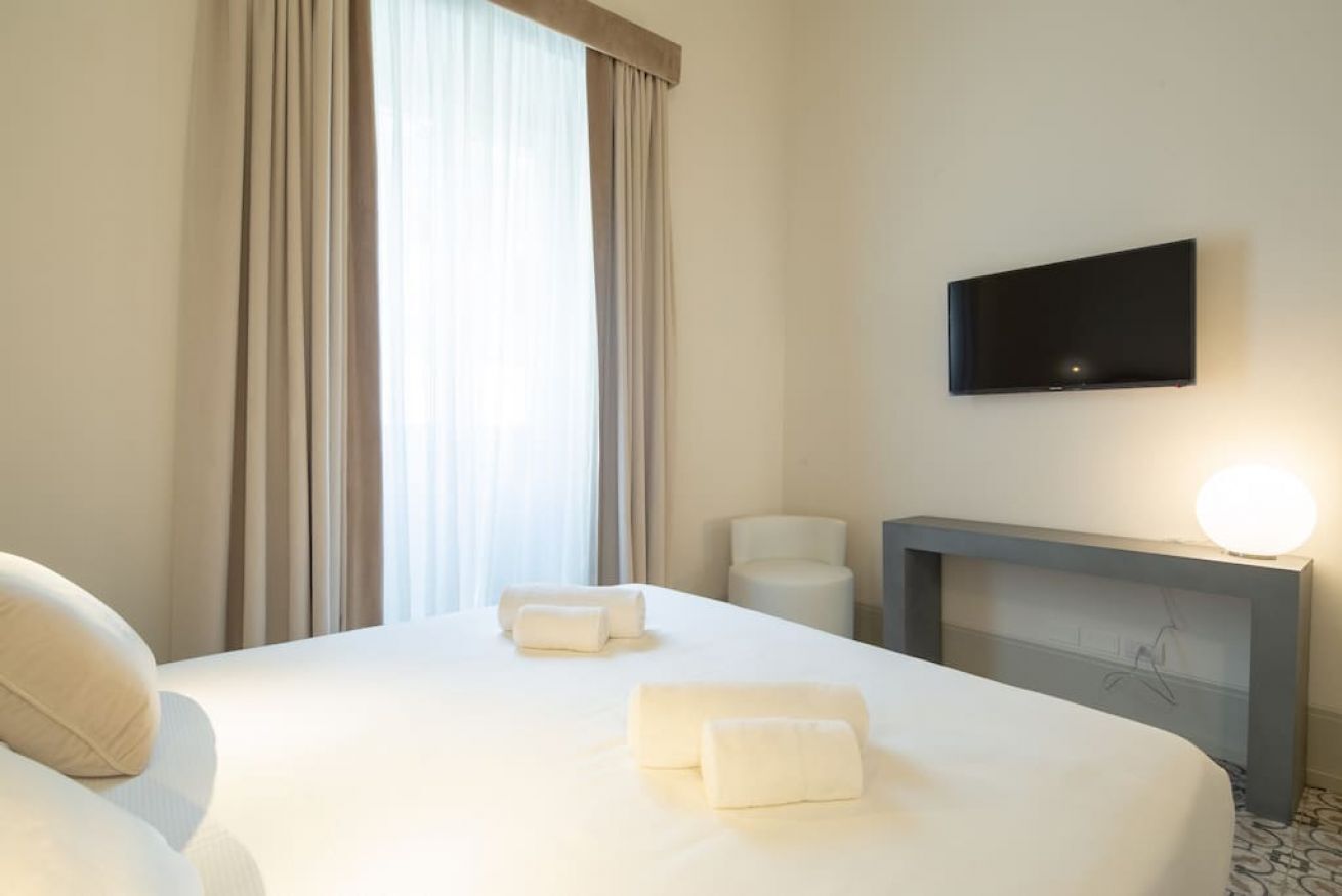 A hotel room with a bed, a flat-screen TV mounted on the wall, a bedside table with a lamp, and a window with curtains.