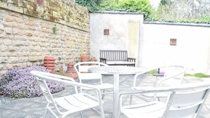 Een terras buiten met een ronde tafel en vier witte stoelen. Achtergrond toont een bakstenen muur met een stenen plaquette en een bank.