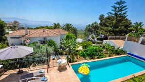 Una piscina con una decoración de piña, sillas de descanso y vistas a una casa y montañas.