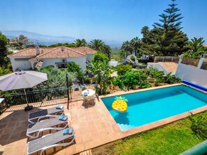 Piscina com decoração de abacaxi, espreguiçadeiras e vista para uma casa e montanhas.