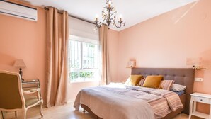 Un dormitorio con una cama grande, una mesita de noche, una silla, una ventana con cortinas y un candelabro.
