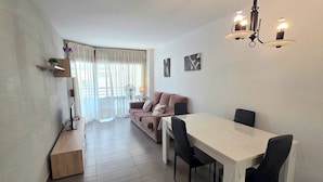A modern living room with a sofa, dining table, and chairs, a small wooden side table, and a ceiling light fixture. - HomeHolidaysRentals Frida - Costa Barcelona (Pineda de Mar)