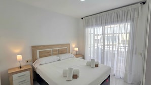 A neatly made bed with white linens, two bedside lamps, a wooden headboard, a nightstand with drawers, and a window with sheer curtains. - HomeHolidaysRentals Frida - Costa Barcelona (Pineda de Mar)