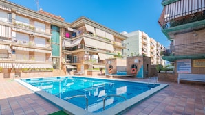 A swimming pool surrounded by multi-story residential buildings with balconies, a small brick structure with a water fountain, and a green mat in the pool area. - HomeHolidaysRentals Frida - Costa Barcelona (Pineda de Mar)
