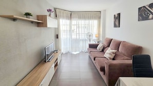 A modern living room with a flat-screen TV, a sofa, a small table, and a window with sheer curtains. - HomeHolidaysRentals Frida - Costa Barcelona (Pineda de Mar)