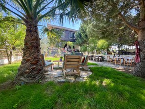 An outdoor seating area with wooden benches and tables, surrounded by lush greenery and a palm tree. - La Kalinda Bozcaada (Bozcaada)