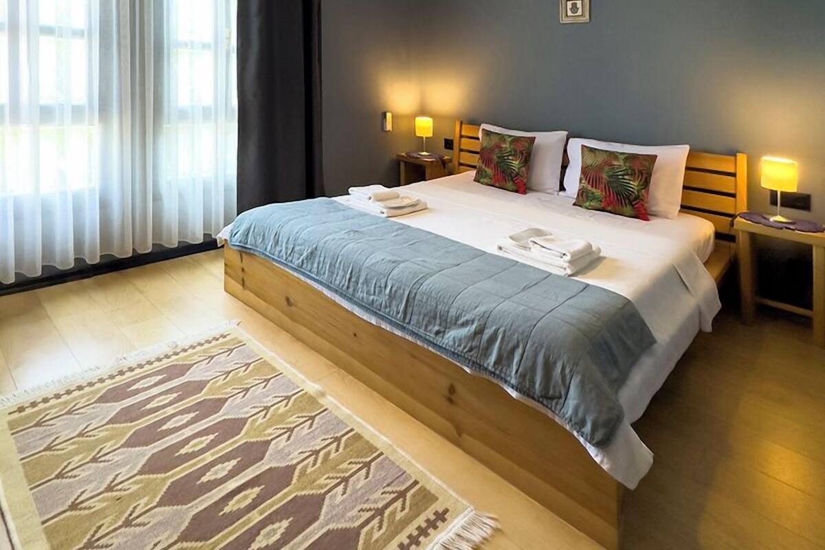 A bedroom with a large bed, two bedside lamps, a patterned rug, and a window with sheer curtains.