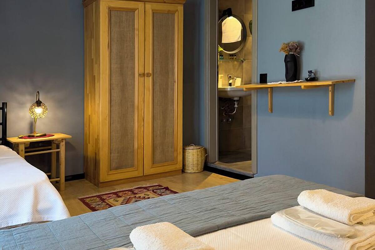 A bedroom with a bed, a wooden wardrobe, a mirror, a shelf with toiletries, and a bedside table with a lamp.