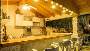 A rustic kitchen with wooden beams, a stone wall, and a bar area with white bar stools. - Etno House Wagner (Peracko Blato)