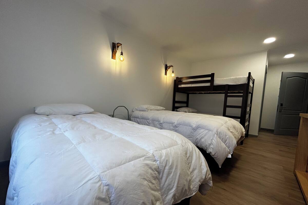 A dormitory room with two bunk beds, a wooden desk, and a wall-mounted light fixture.