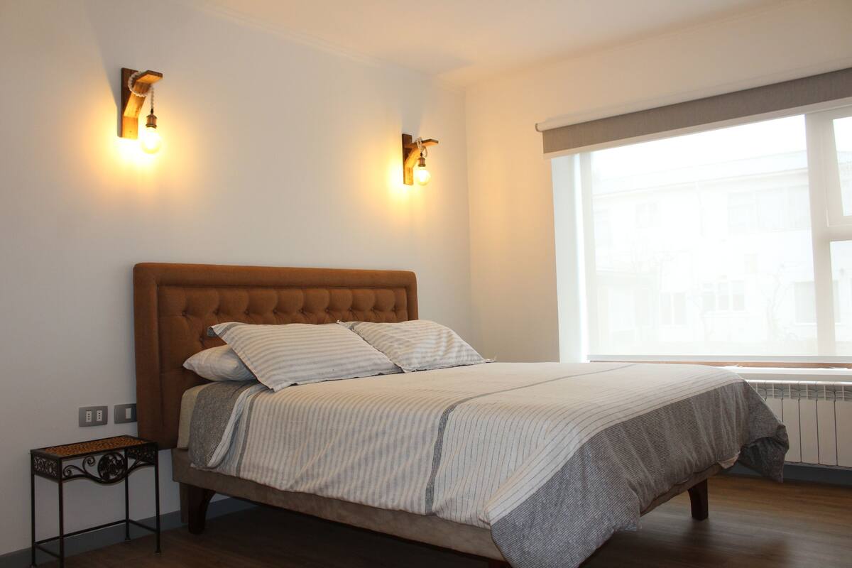 A bedroom with a large bed, two wall-mounted lights, a small side table, and a window with a view of buildings.