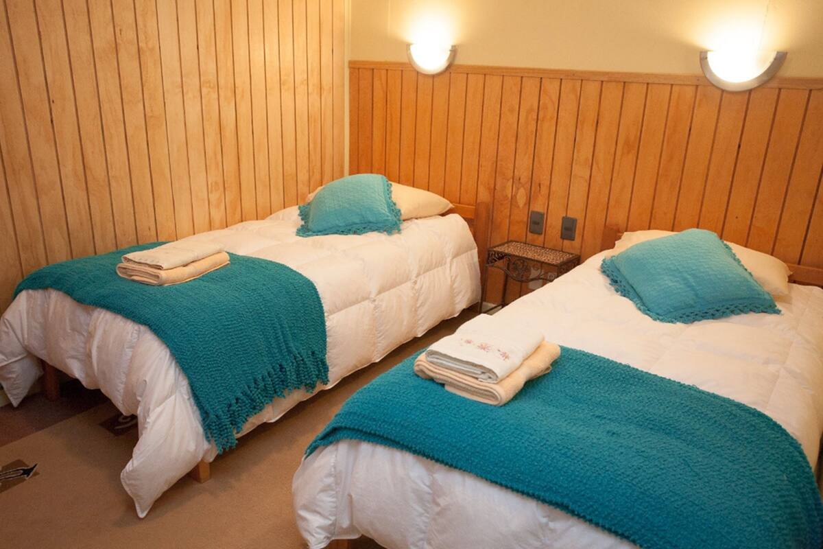 Two twin beds with teal blankets and white linens, wooden paneling on the walls, and a small nightstand between them.