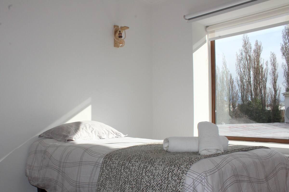 A bedroom with a bed, a window with a view of trees, and a wall-mounted light fixture.