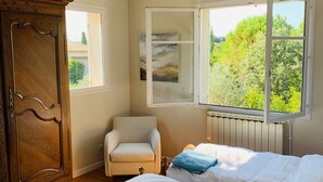 A bedroom with two beds, a wooden wardrobe, a chair, and a window with a view of greenery.