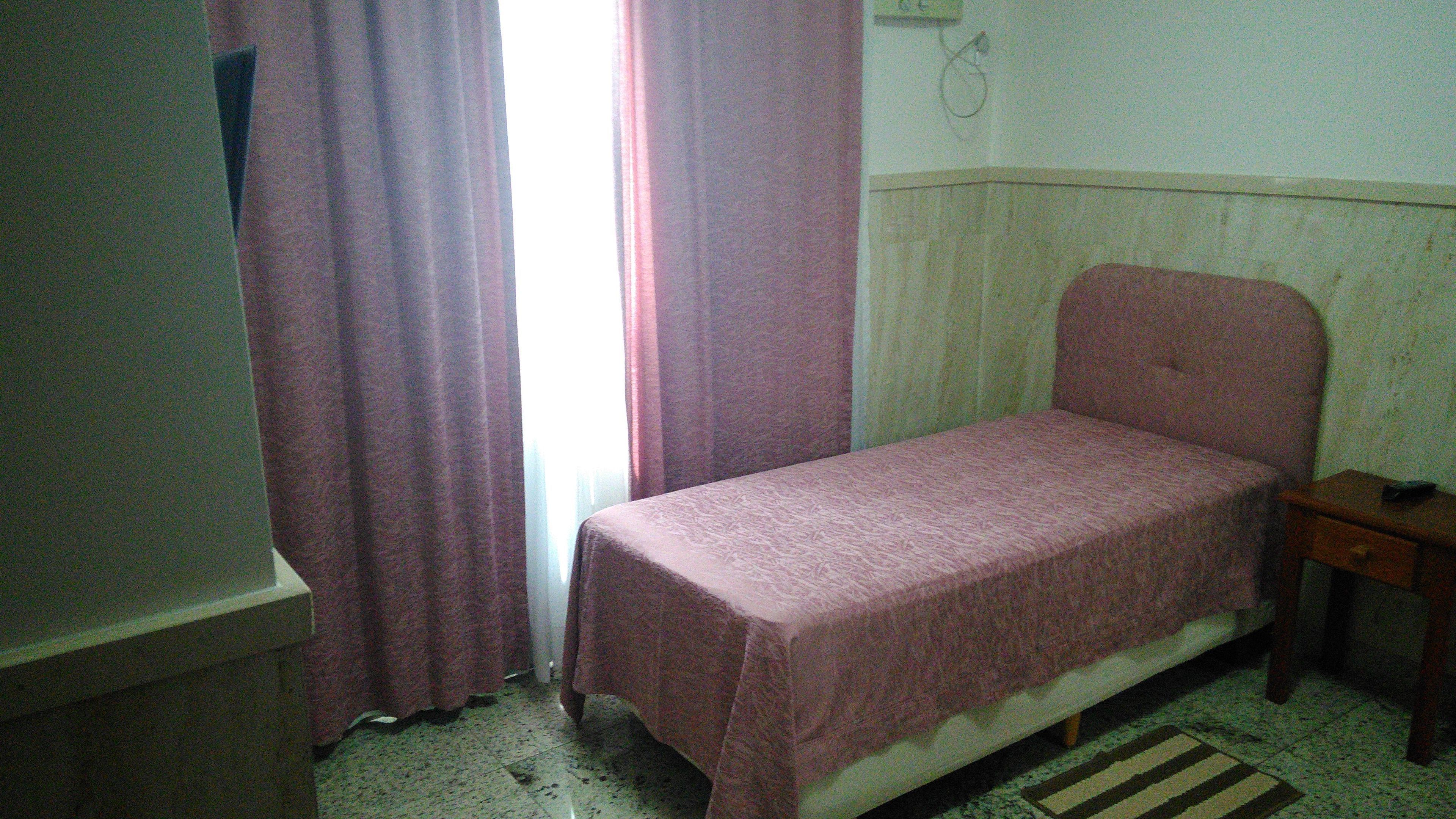 A small, single-bed room with a plain bed, a bedside table, and a window with curtains.