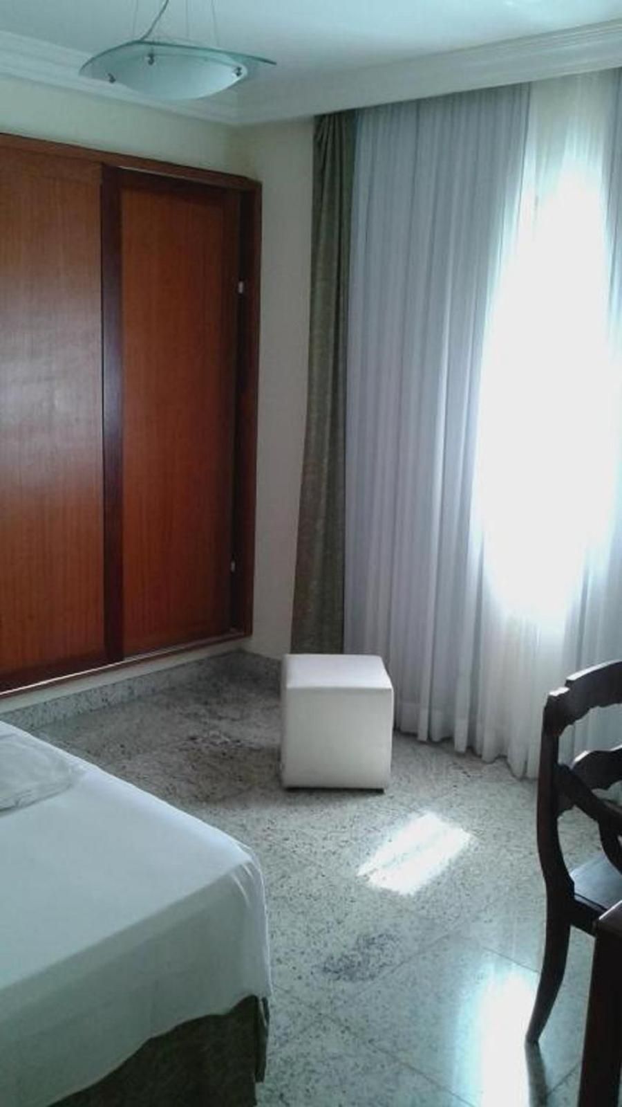 Una habitación de hotel con cama, un otomán blanco pequeño, un armario de madera y un ventanal con cortinas.
