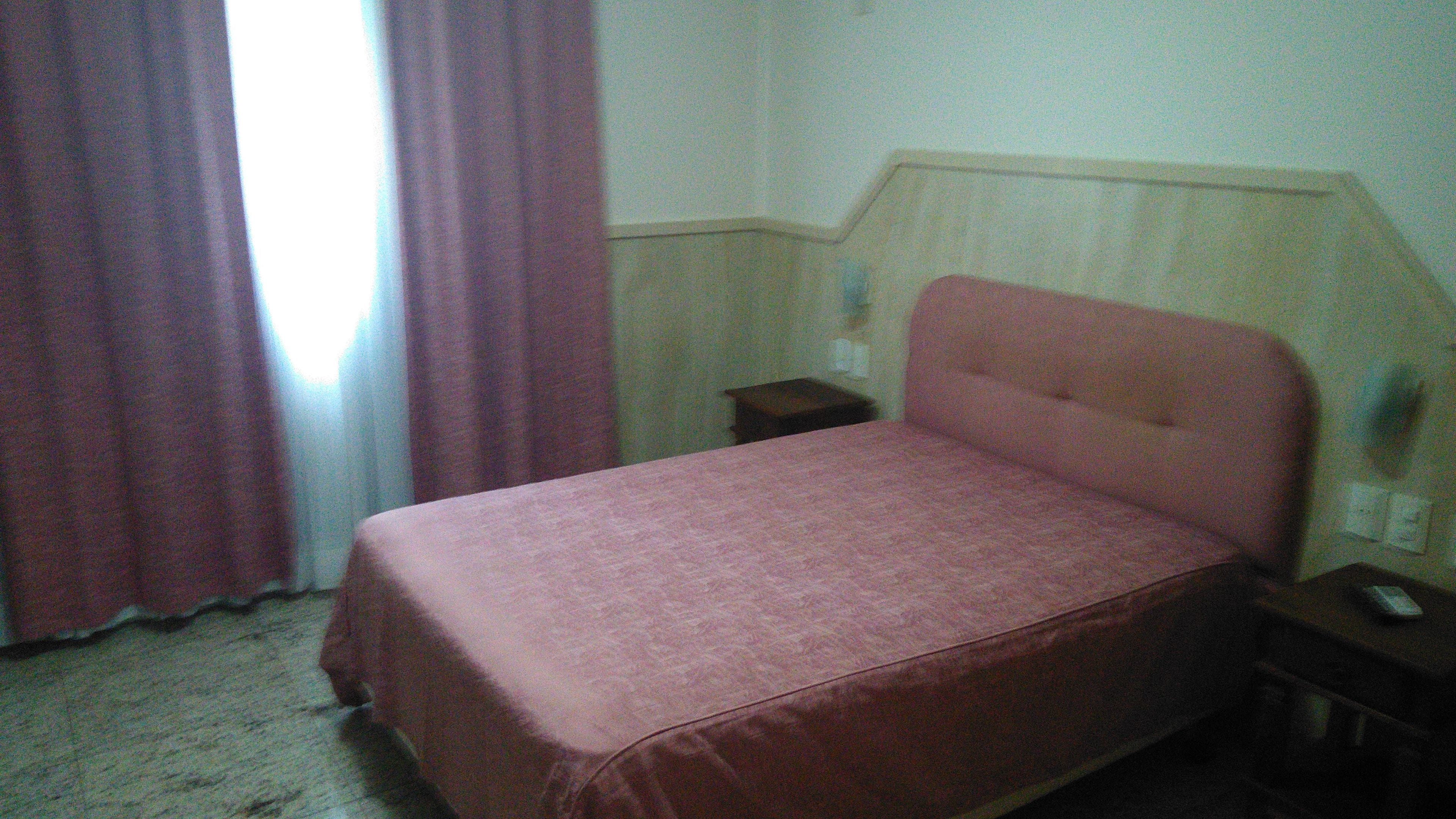 Una cama individual con cabecera, mesita de noche, lámpara y ventana con cortinas.