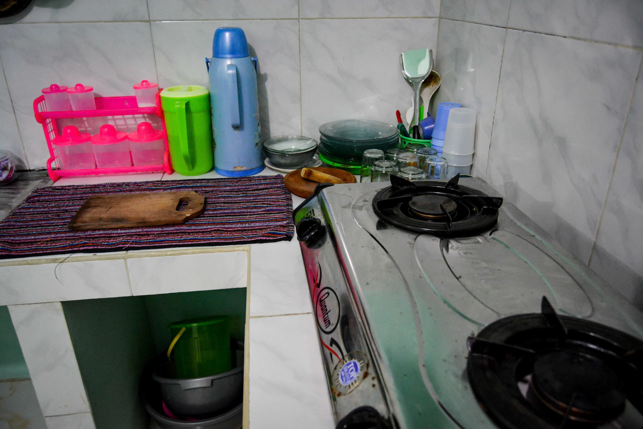 Una cocina con estufa a gas, tabla de cortar, pileta y diversos utensilios y recipientes de cocina.