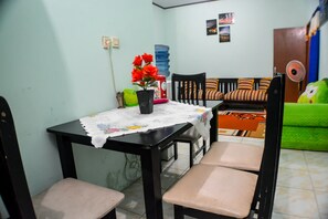 Un comedor con una mesa, sillas y un banco. Hay una cesta verde, un jarrón con flores y un ventilador en la pared.