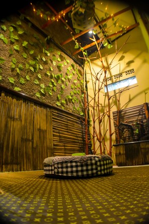 A room with a checkered cushion on a patterned bedspread, a bamboo wall, and a window with a view.