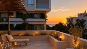 A modern outdoor seating area with a sofa and chairs, surrounded by plants and a sunset view. - Welcome to Lumen Villa the first of the Adelos Trilogy (Elliniko)