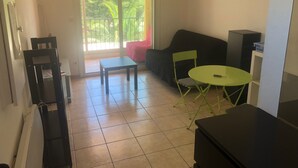 Un salon avec un canapé, une petite table et un fauteuil vert. Une porte-fenêtre coulissante donne accès à un balcon d’où l’on peut admirer la vue sur les arbres.