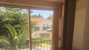 Une pièce avec un fauteuil rouge, une porte-fenêtre coulissante donnant sur un balcon avec vue sur un quartier résidentiel, et un sol carrelé.