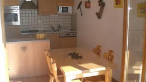 Eine Küche mit Holzausstattung, einer Mikrowelle und einem Spülbecken. Ein Esstisch mit Stühlen und ein Fenster mit Vorhängen sind ebenfalls vorhanden.
