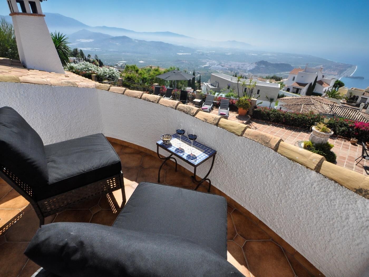 Une terrasse avec deux chaises noires dotées de coussins, une petite table avec un verre, et une vue sur la mer et les montagnes.