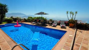 A swimming pool with a view of the ocean and mountains, surrounded by lounge chairs and potted plants.