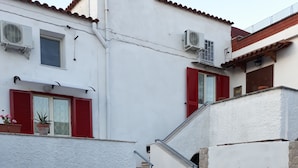 Ein Innenhof mit einer Treppe, die zu einem zweiten Stock führt, umgeben von weißen Wänden und roten Fensterläden, mit Blumentöpfen und einem kleinen Zaun.