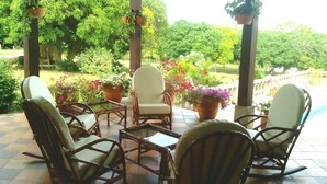 Uma área de estar coberta ao ar livre com cadeiras e mesas de vime, rodeada por plantas em vasos e vista para um jardim exuberante.