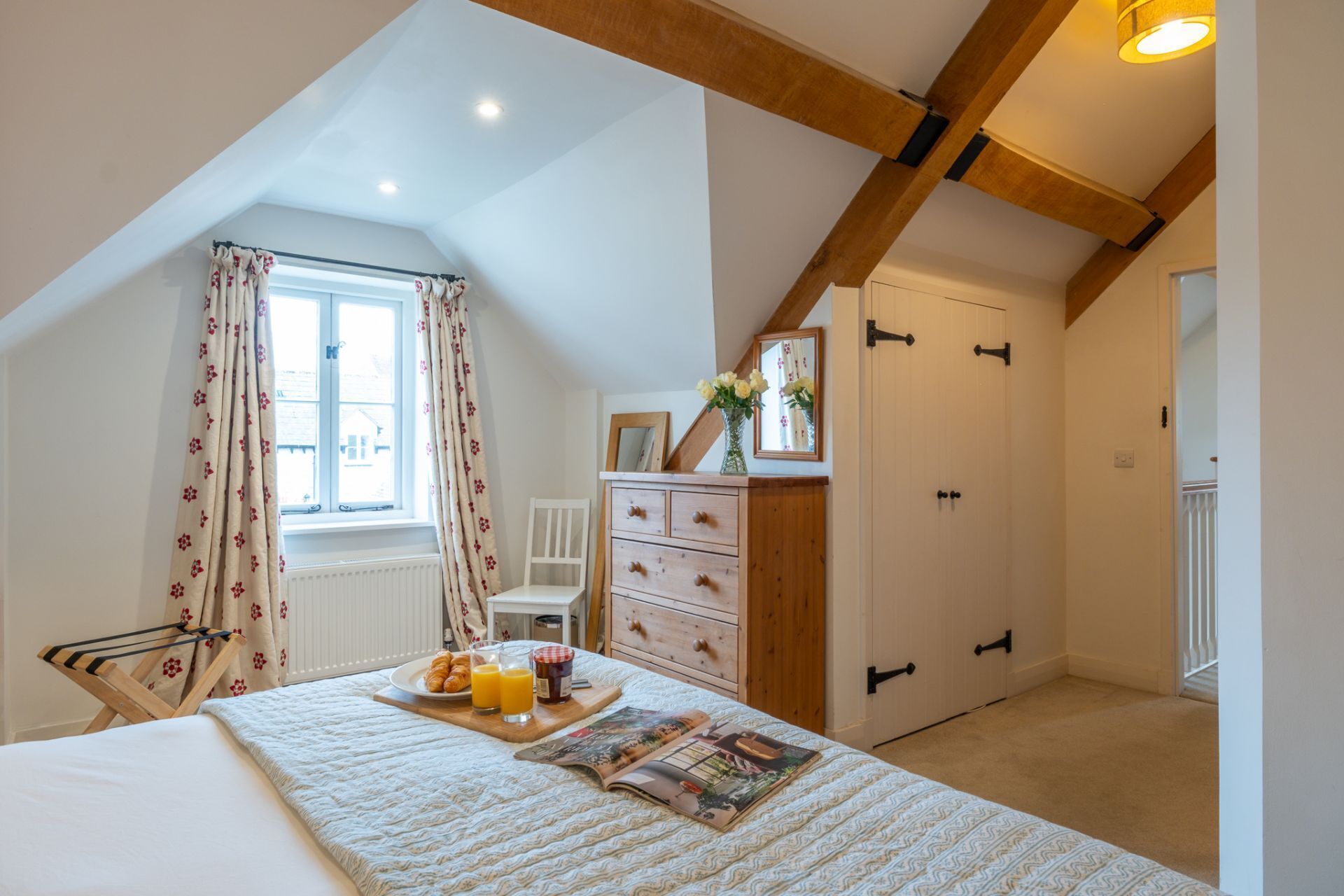 Un dormitorio con techo inclinado, una cama con una revista y alimentos para el desayuno, un aparador y una ventana con cortinas.
