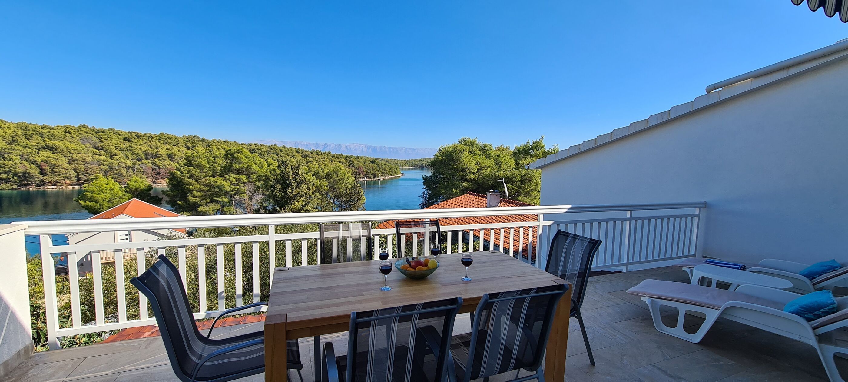 A balcony with a wooden table, chairs, and a view of a lake and forest.